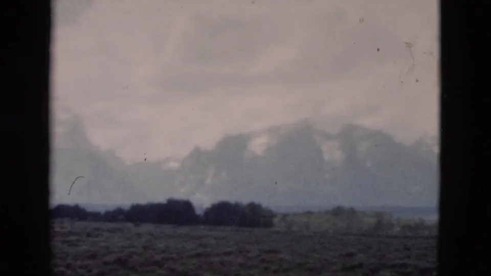 1958 8mm view of the Grand Teton range from the Yellowstone, Wyoming metadata area, framed by dark vertical window edges over a muted plain.