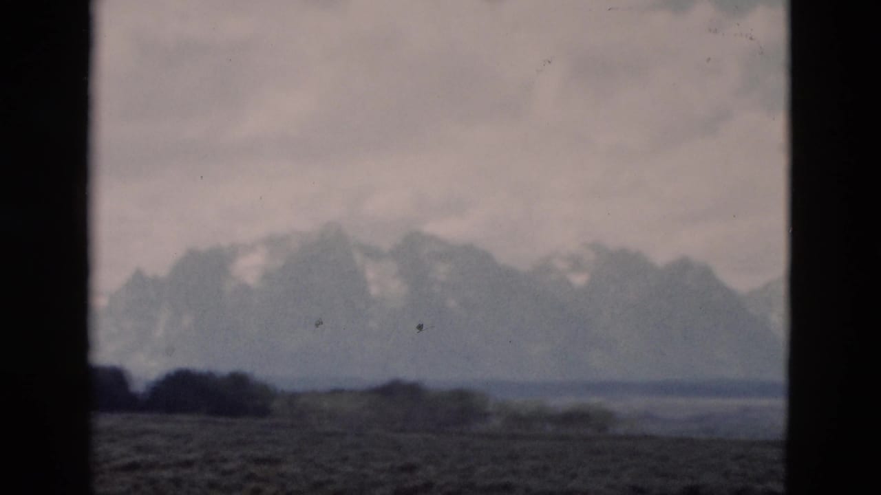 Hazy mountain peaks and low trees viewed through dark window dividers under a cloudy sky.
