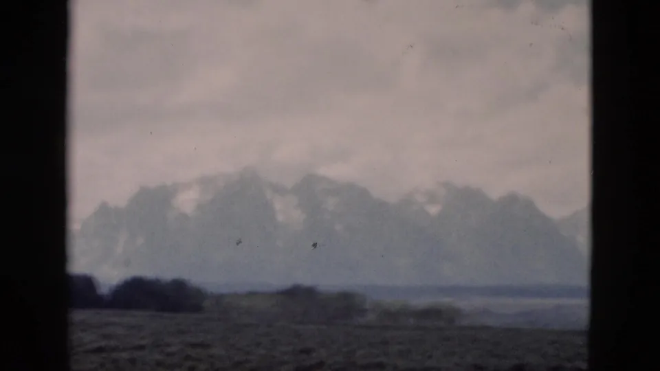 Grand Teton mountain scenery appears in soft focus through dark window framing, with an open foreground plain and overcast sky.