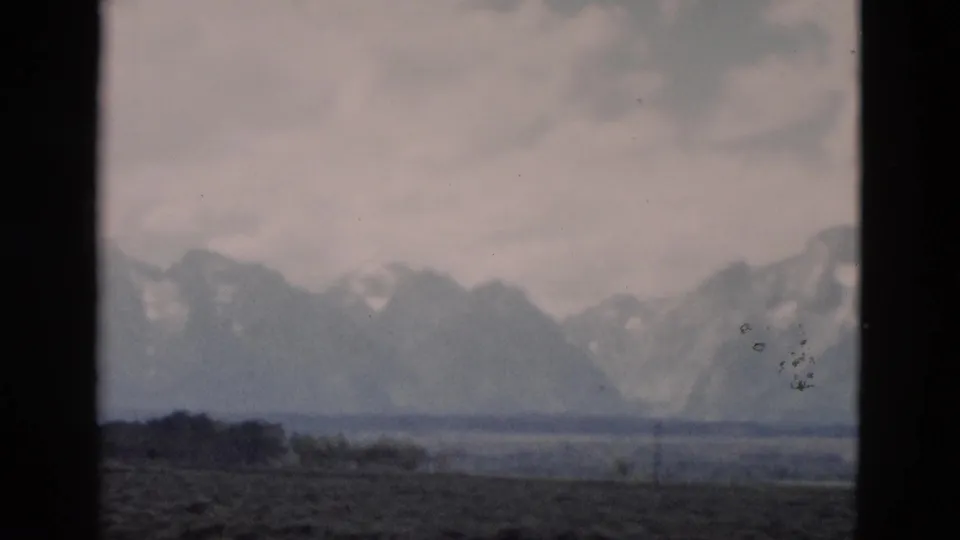 A 1958 visitor-shot 8mm frame shows the Grand Teton range beyond open land, with dark film-window borders and visible dust marks.