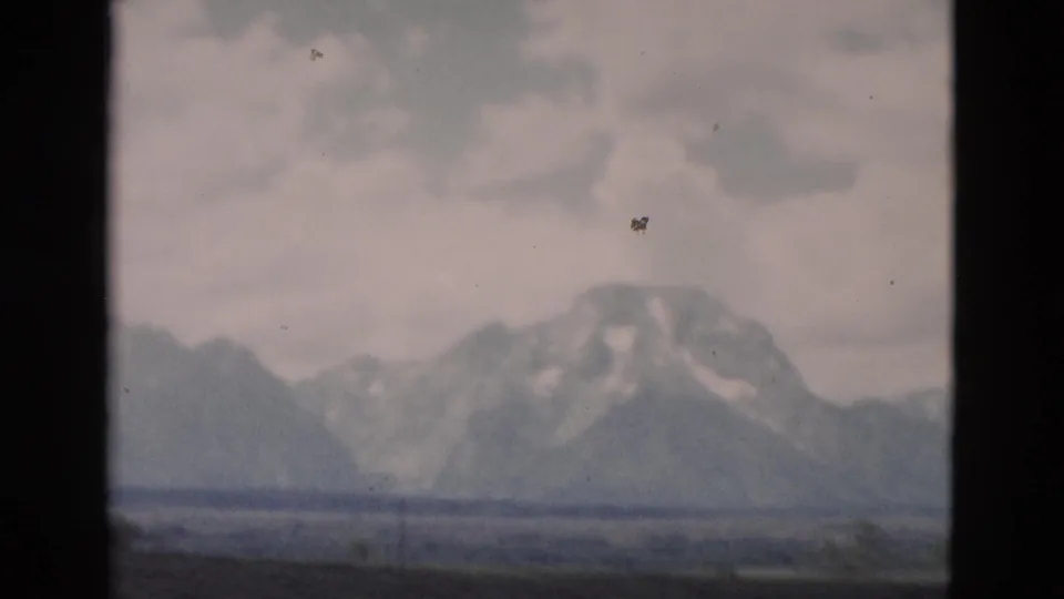 The final 8mm frame holds on a snow-marked Grand Teton peak beyond open ground and low vegetation, with soft focus and visible film wear.