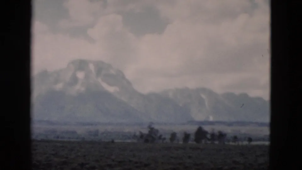 1958 Yellowstone home-movie frame with a snow-marked mountain slope, gray clouds, scattered trees, and visible 8mm grain.