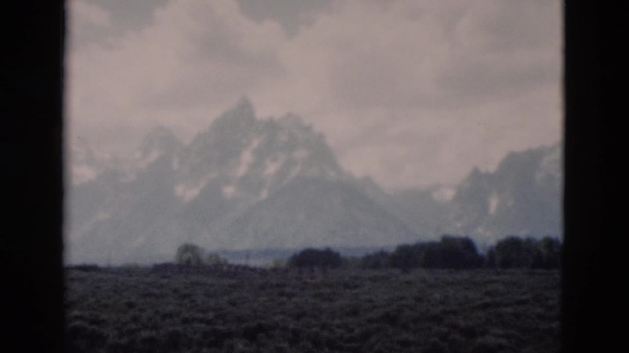 1958 8mm Yellowstone travel frame showing distant snow-streaked mountains beyond a dark open meadow under clouds.
