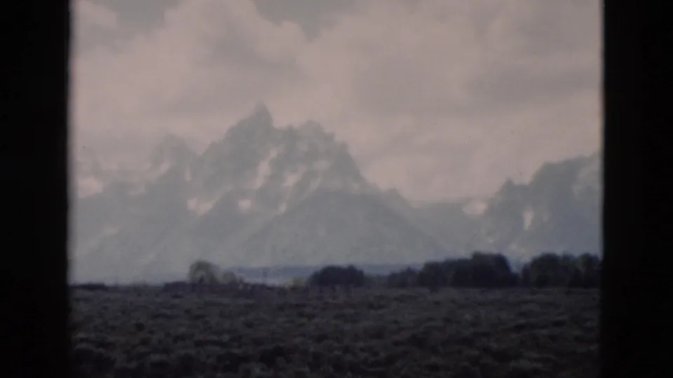 1958 8mm Yellowstone travel frame showing distant snow-streaked mountains beyond a dark open meadow under clouds.