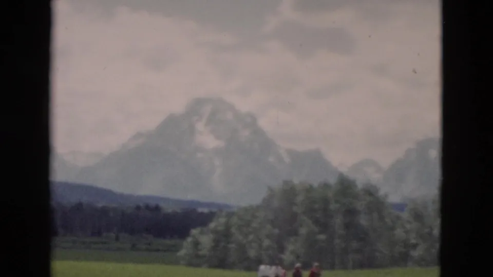 1958 Yellowstone home-movie frame with mountains, trees, and small visitors visible near the bottom edge.
