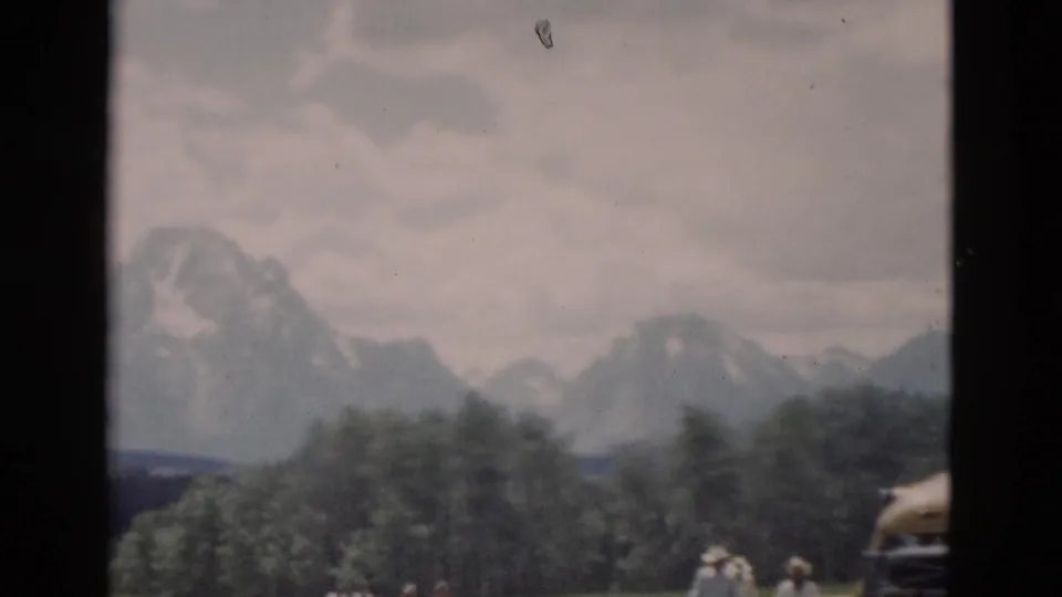 1958 Yellowstone travel frame showing trees before distant mountains, with people and part of a yellow car at the right edge.