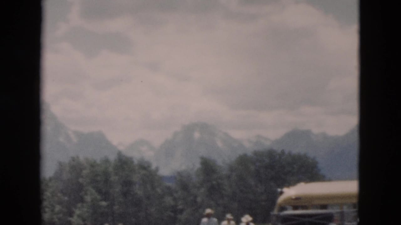 1958 8mm home-movie frame showing a yellow excursion bus, small visitors, trees, and distant snow-capped mountains in Wyoming park scenery.
