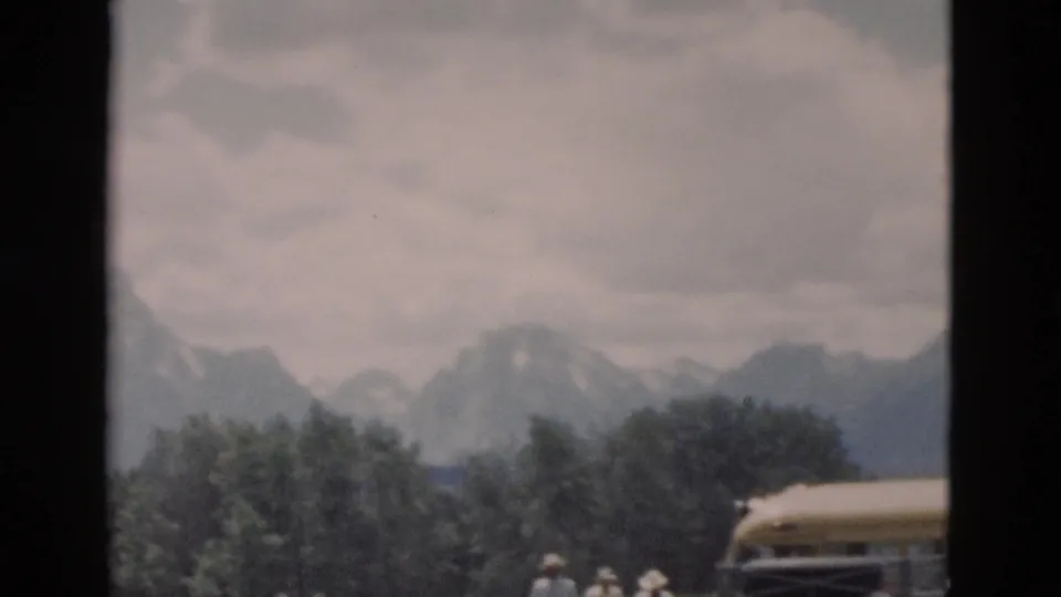 1958 8mm home-movie frame showing a yellow excursion bus, small visitors, trees, and distant snow-capped mountains in Wyoming park scenery.