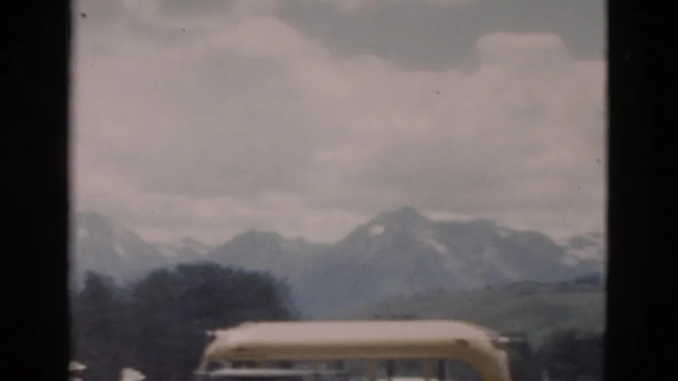 Yellow excursion bus partly visible in the foreground, with green terrain and a snow-capped mountain range under cloudy sky.