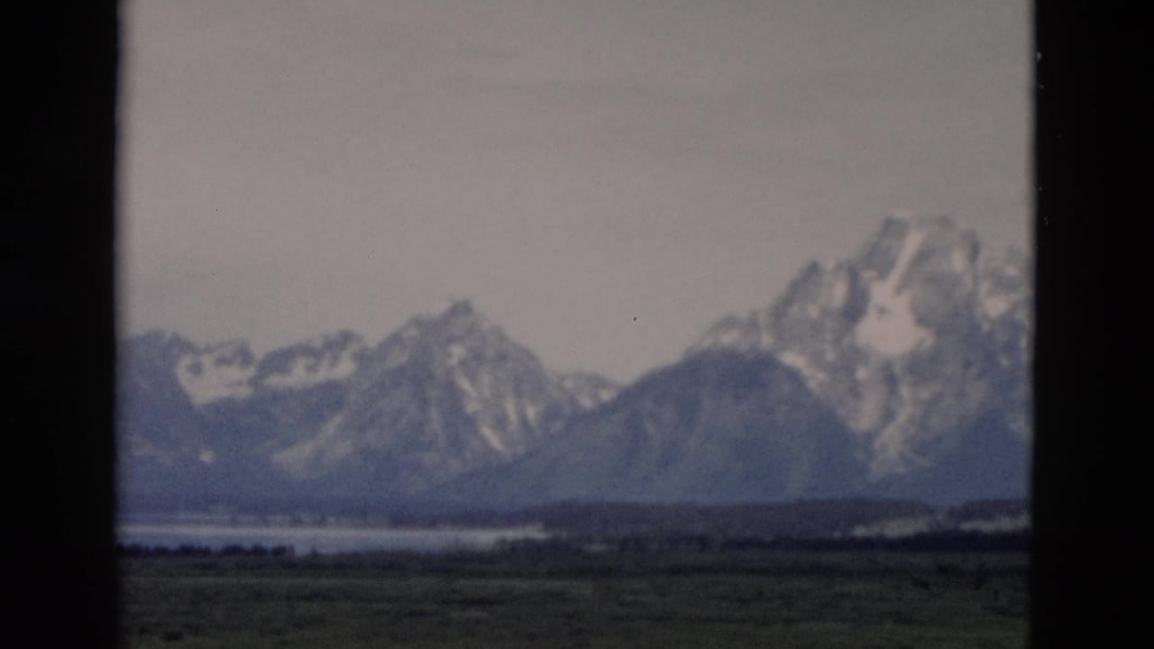 1958 8mm Yellowstone, Wyoming view of distant snow-covered mountains, green fields, and water framed by dark film edges.