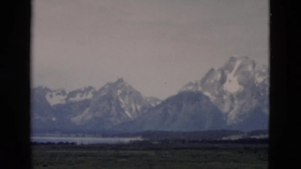 1958 8mm Yellowstone, Wyoming view of distant snow-covered mountains, green fields, and water framed by dark film edges.
