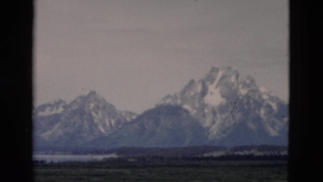 1958 8mm Wyoming mountain landscape with snow-streaked peaks, muted fields, water, cloudy sky, and prominent film gate border.