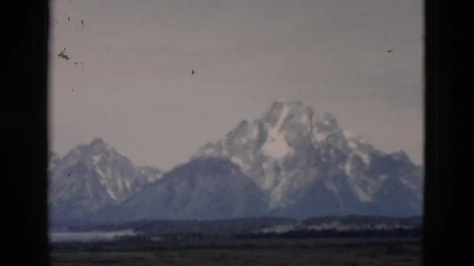 1958 8mm Yellowstone, Wyoming landscape showing a large snow-covered peak, surrounding ridges, green fields, and a low waterline.
