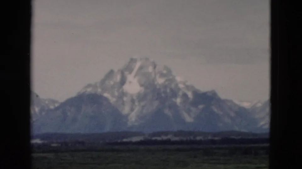 1958 8mm Wyoming view of a snow-covered mountain and foothills across green fields, with soft focus and dark film borders.