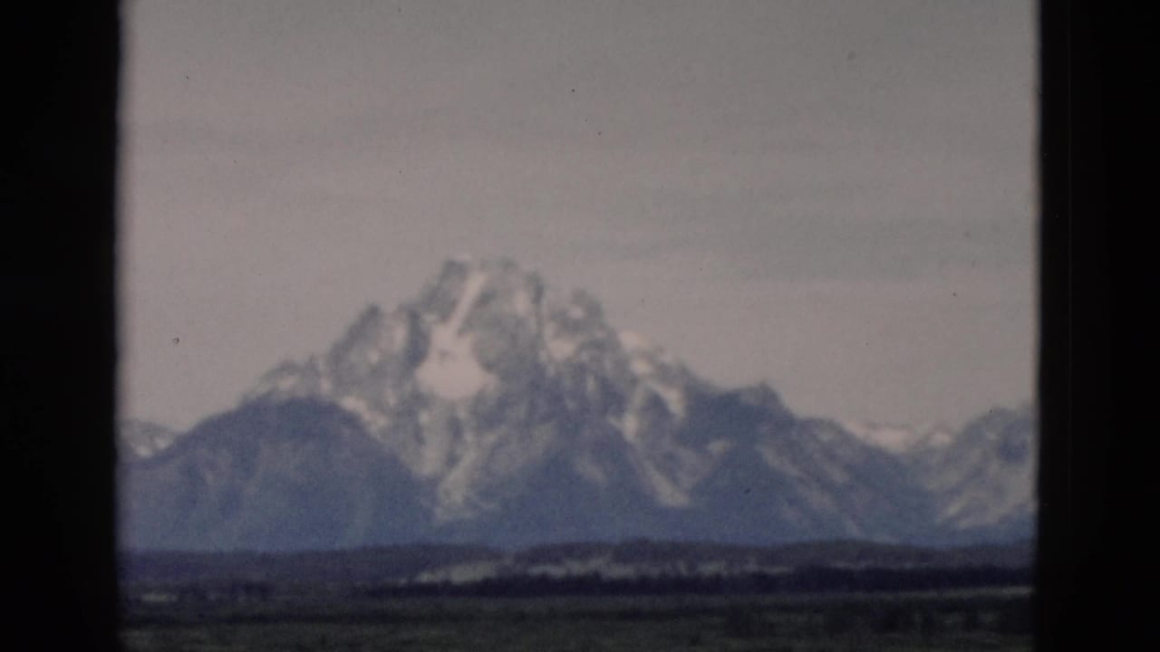 Snow-capped mountain range seen beyond dark 8mm frame borders and a green foreground.