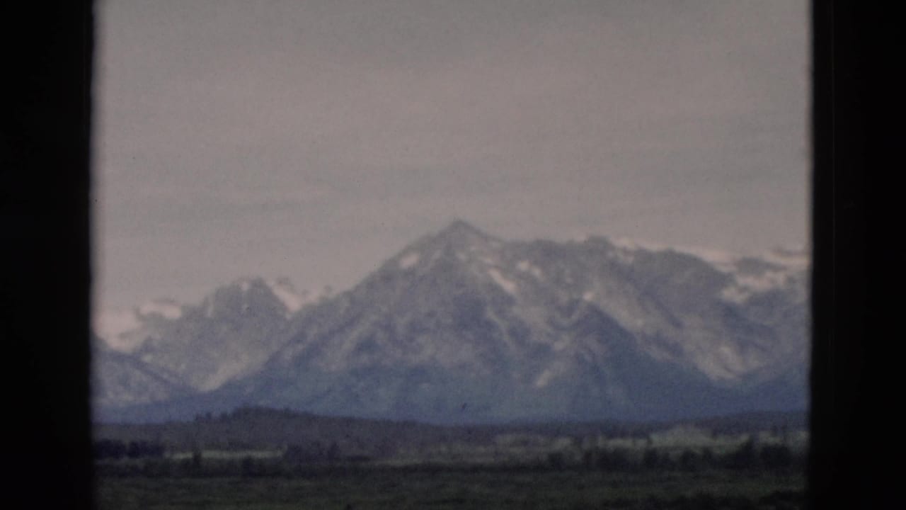 Snow-streaked mountain range beyond a green valley, seen through dark vertical framing.