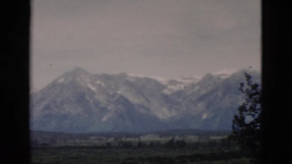 1958 Yellowstone-area mountain view with snow-streaked peaks, open valley and a dark tree silhouette at right.