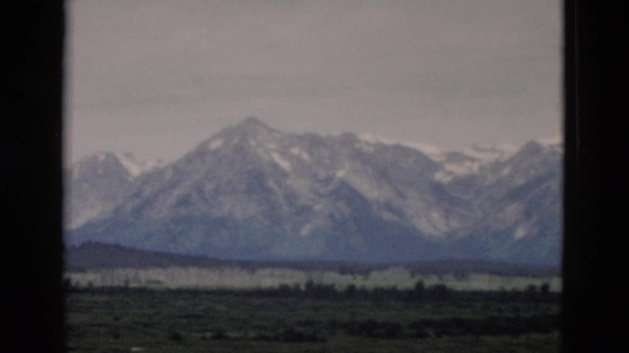 1958 Yellowstone-area 8mm frame showing distant snow-streaked mountains above a green valley under a hazy sky.