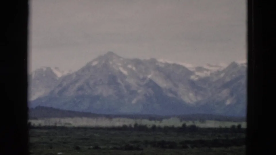 1958 Yellowstone-area 8mm frame showing distant snow-streaked mountains above a green valley under a hazy sky.