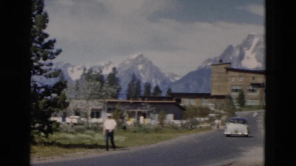 1958 Yellowstone-area roadside view with a stone lodge-like building, mountain backdrop, pale car, and one visible man.