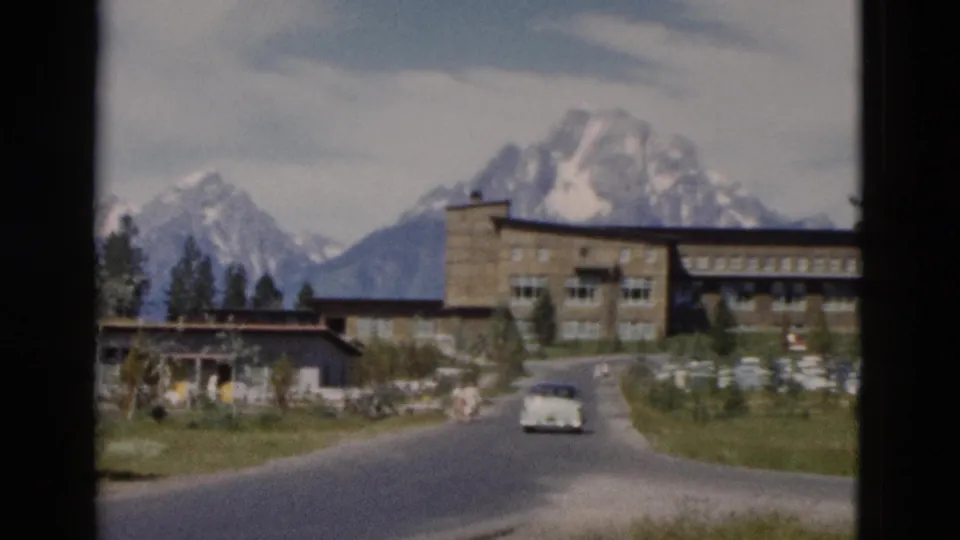 Mid-century road approach in the Yellowstone area, showing a pale car, large stone lodge-like building, trees, and mountains.