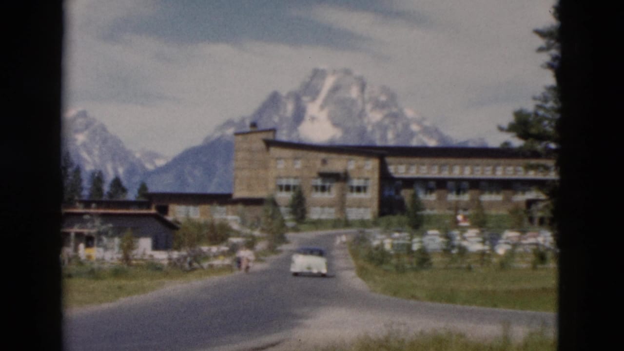 Stone building beside a curved road with cars, lawn, trees, and snow-capped mountains beyond.