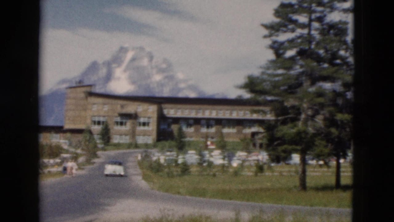 1958 Yellowstone frame with a large stone building, road, small distant visitors, parked cars, conifers, and snow-covered peaks.