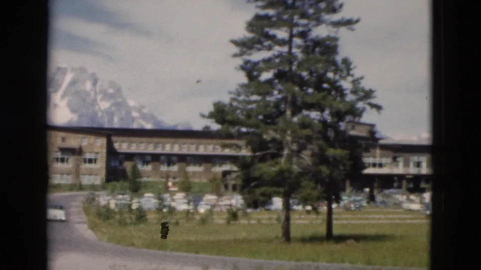 1958 Yellowstone home-movie view with a stone building partially obscured by trees, road edge, parked cars, lawn, and mountain scenery.