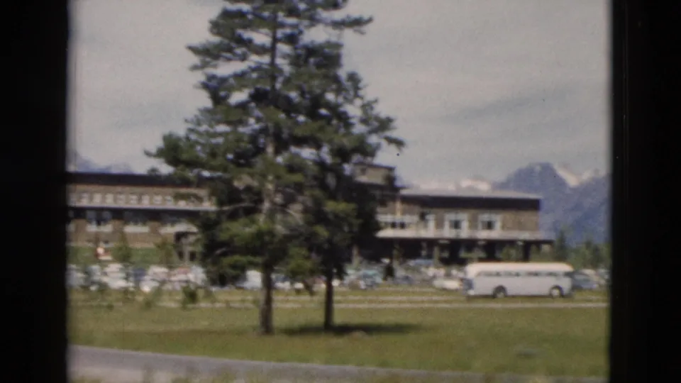 1958 Yellowstone frame showing conifers in front of a stone building, parked vehicles, a white bus-like vehicle, grass, and distant peaks.