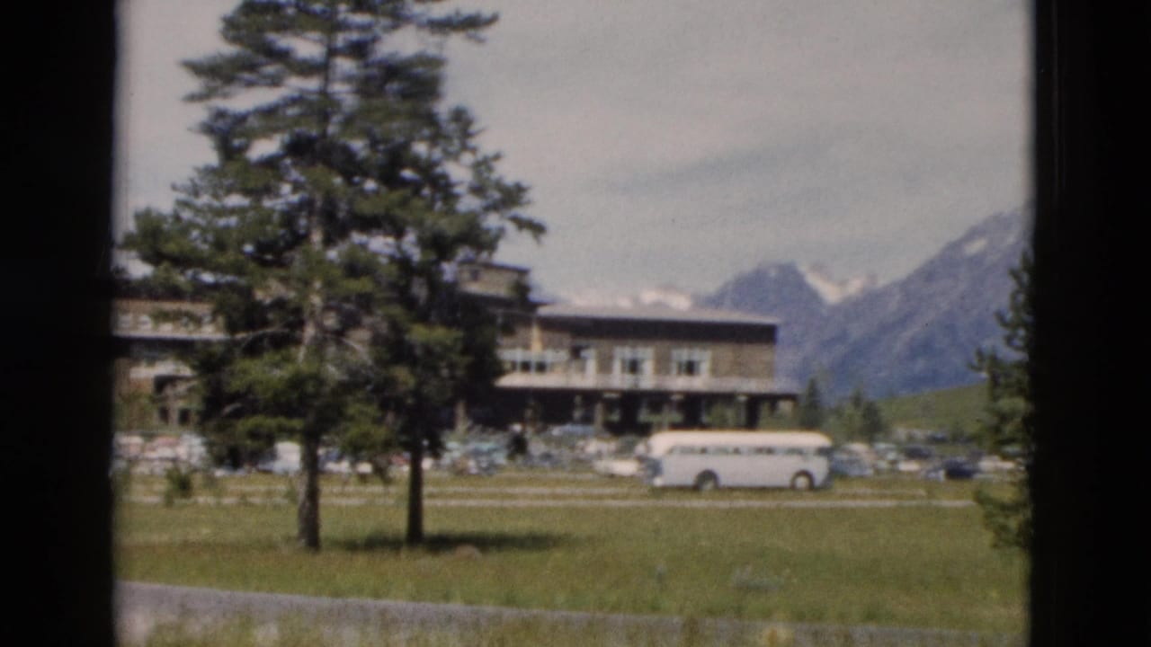 Lodge-style building with a white bus, lawn, evergreens, and mountains behind it.