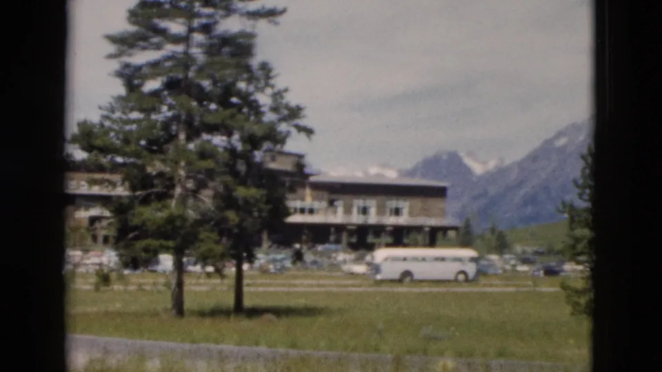 1958 Yellowstone-area 8mm frame showing a lodge-style building, white tour bus, grassy lawn, evergreens, and distant mountains.