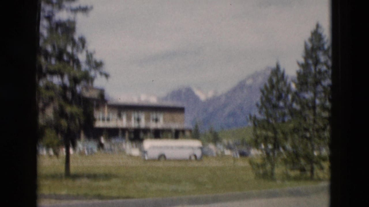1958 Yellowstone-area view of a white bus near a lodge or motel building, framed by trees and a mountain backdrop.