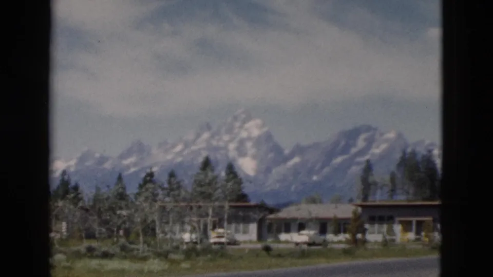 1958 Yellowstone-area 8mm frame showing roadside cabins or motel units, parked cars, evergreens, and snow-capped mountains.