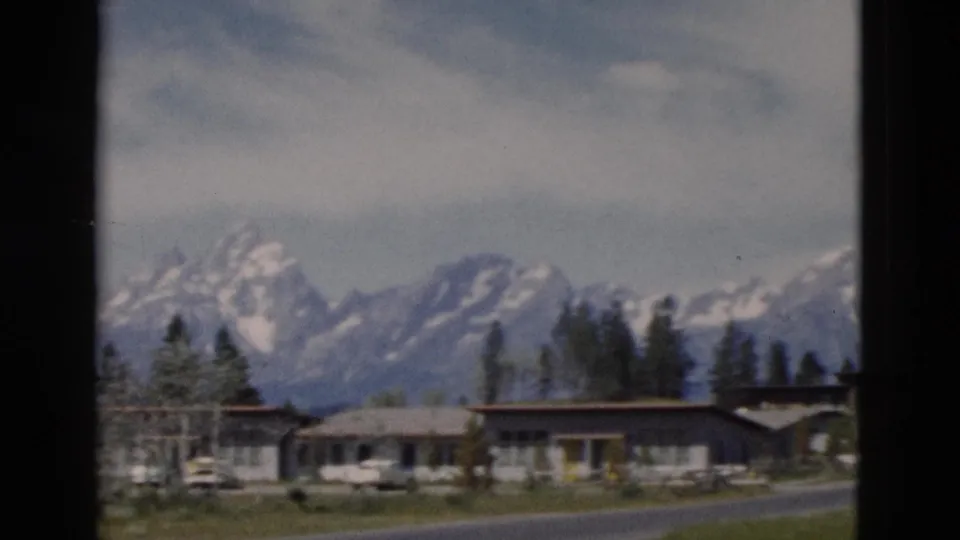 1958 Yellowstone-area home-movie frame of low lodging buildings, parked cars, trees, road, and a snow-capped mountain range.
