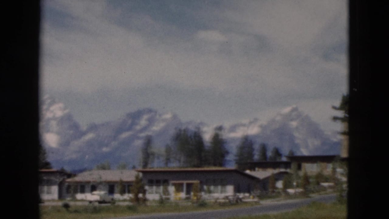 Snowy mountains behind low buildings, road, grass, and dark window-frame borders.