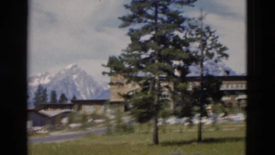 1958 8mm travel frame showing pine trees, lodge buildings, a road, and snow-capped mountains in Yellowstone, Wyoming.