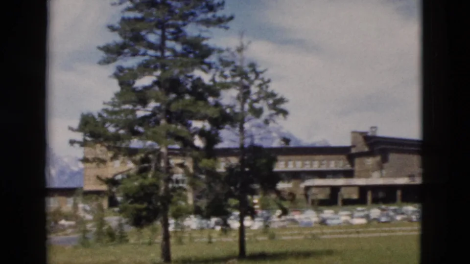 Yellowstone home-movie frame showing a larger lodge building, parked cars, tall pines, grass, and distant snowy peaks.