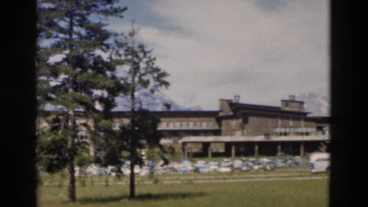 1958 Yellowstone-area lodge exterior seen across grass, with trees, a parking area, and distant snow-capped mountains.