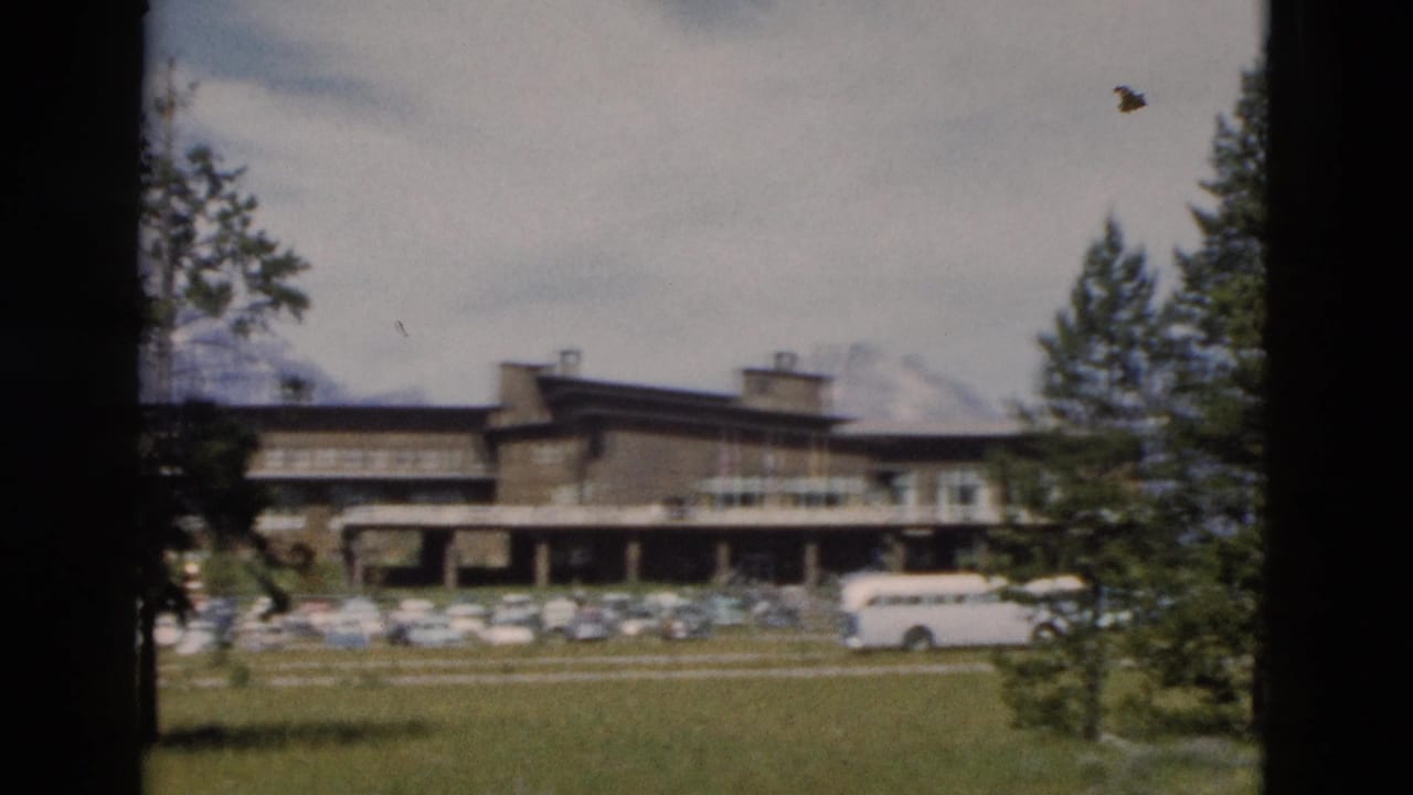 Lodge exterior with a white tour bus, parked cars, trees, grass, and mountains beyond.