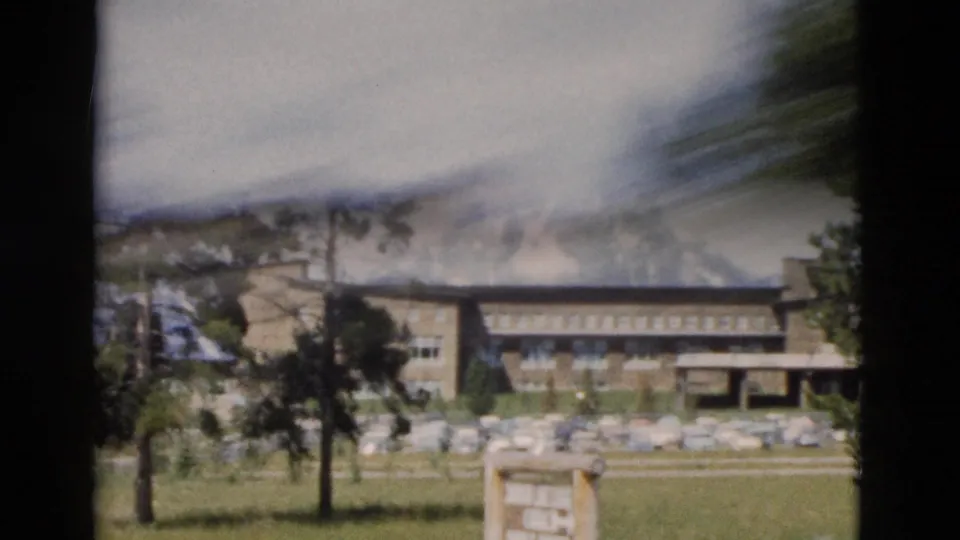 During a handheld pan, the 1958 Yellowstone lodge view softens, with trees, parked cars, grass, and mountain shapes still visible.