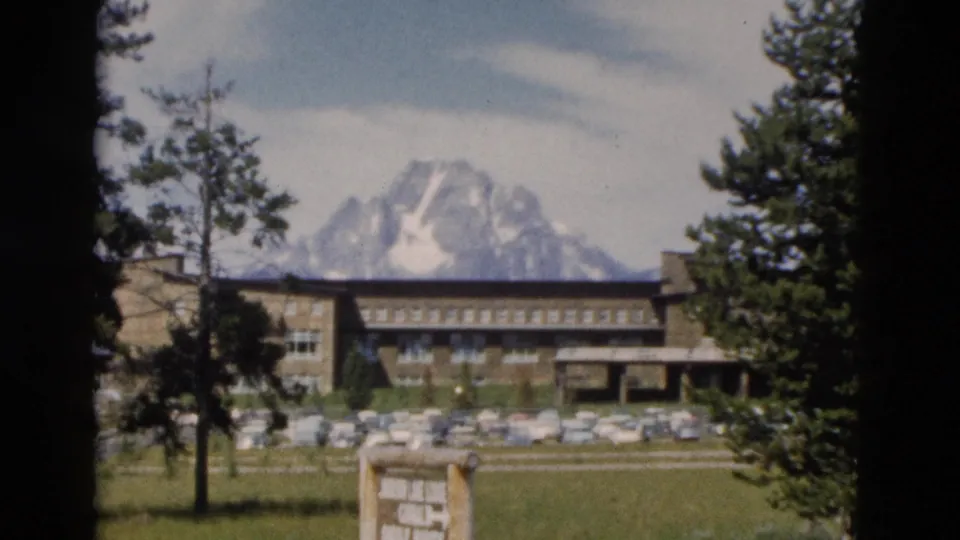 Yellowstone-area lodge in 1958 with classic cars, evergreen trees, open grass, and a snow-capped mountain rising behind it.