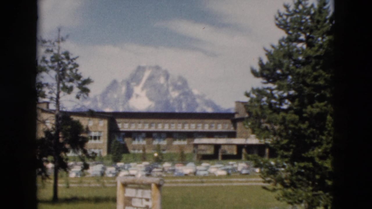 1958 8mm Yellowstone-area travel view with a lodge building, parked cars, trees, and distant snow-capped Teton peaks.
