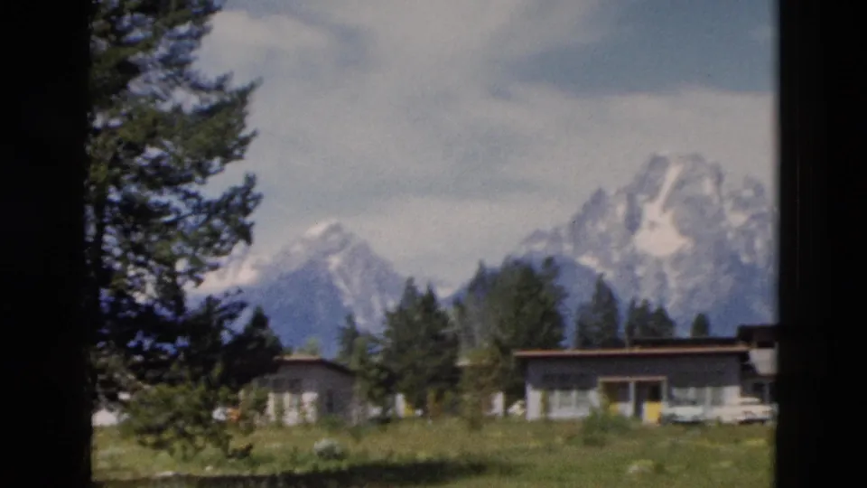 Yellowstone-area 1958 home-movie frame showing Teton peaks behind trees and low visitor buildings, partly obscured by a foreground trunk.