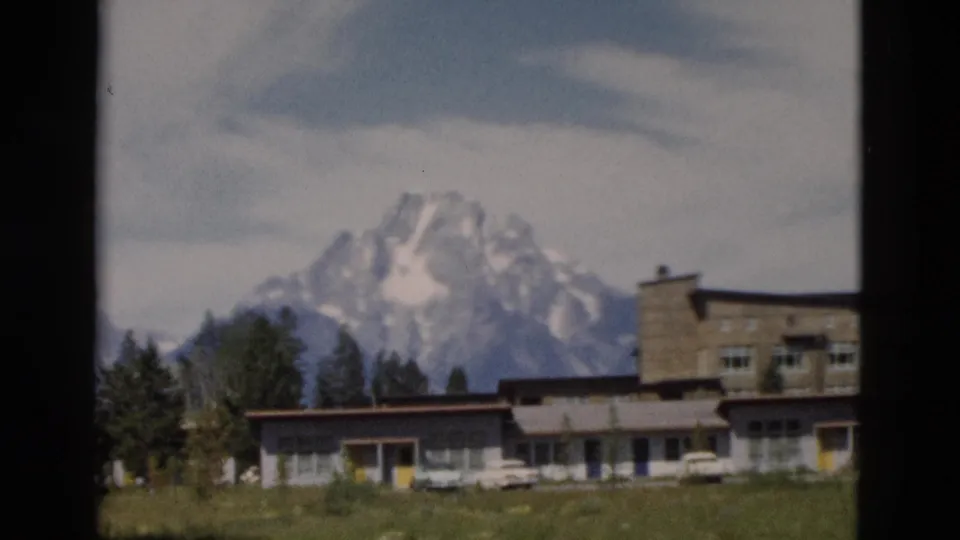 Yellowstone-area 1958 8mm frame showing a snow-capped Teton peak behind trees, low buildings, and part of a larger structure.