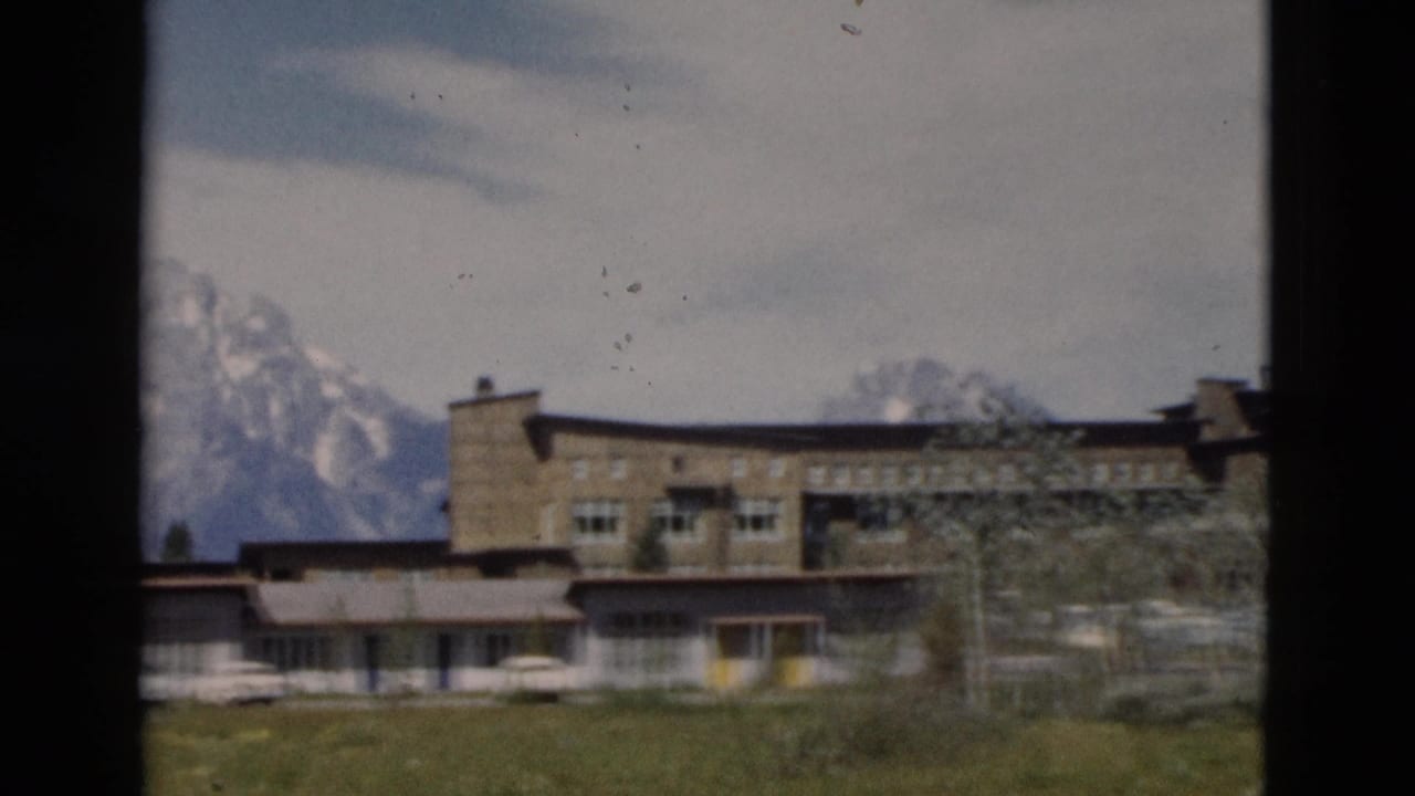 1958 Wyoming frame with a long low-rise structure, pale cars, scattered trees, and a mountain ridge beyond the building.