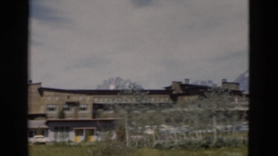 Soft 8mm frame of the Yellowstone-area building complex seen through trees, with grass, parked cars, and distant mountains.
