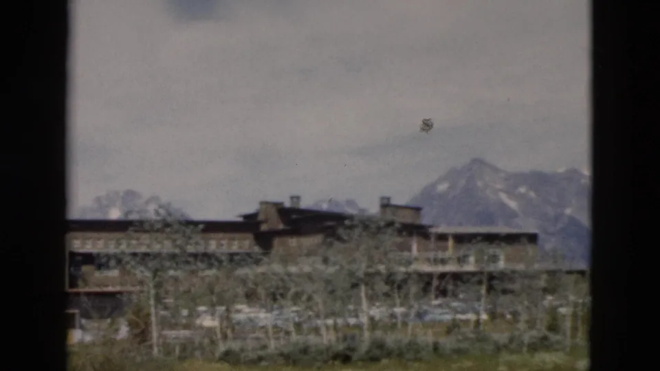 1958 Wyoming 8mm frame showing the end of a long building, bare trees, grassy ground, and a mountain peak to the right.