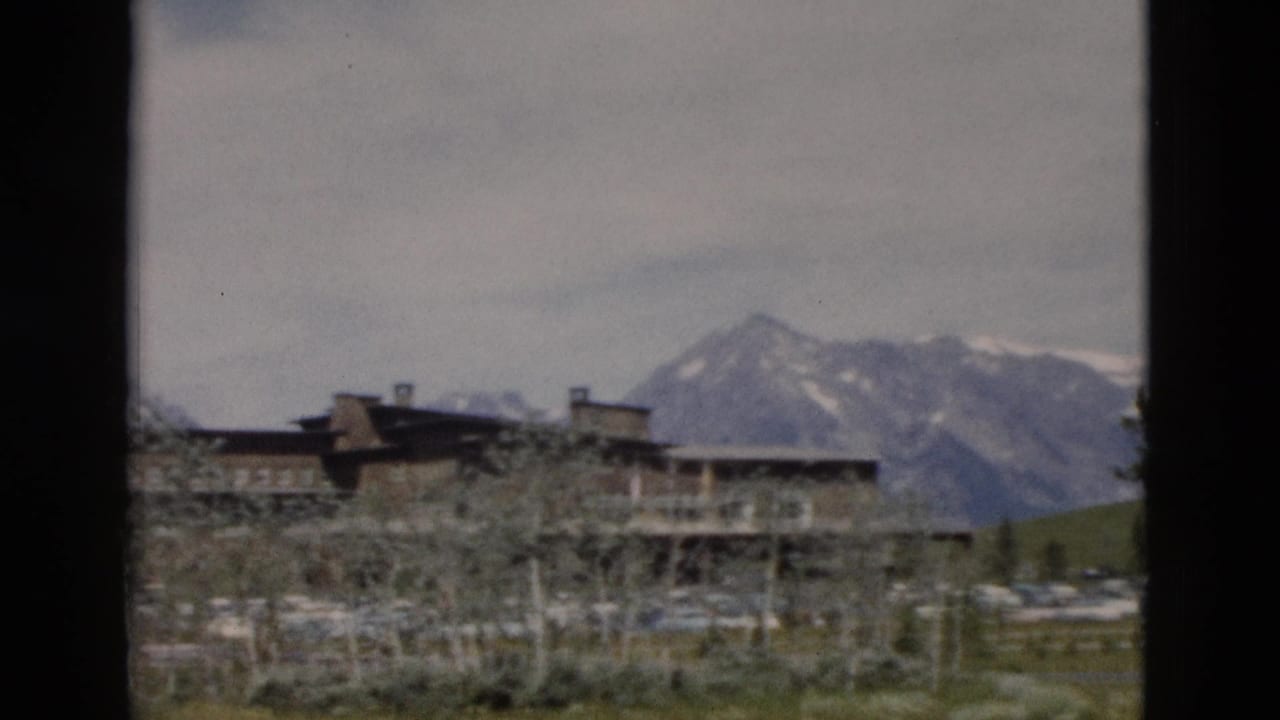 1958 Yellowstone home-movie frame showing a rustic lodge below snow-streaked mountains with trees, grass, and parked cars.