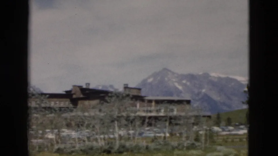 1958 Yellowstone home-movie frame showing a rustic lodge below snow-streaked mountains with trees, grass, and parked cars.