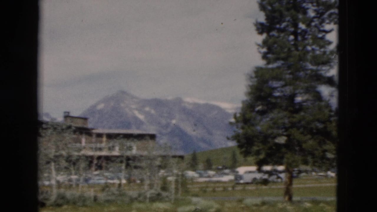 Lodge and distant mountains framed by a tall evergreen and dark vertical film edges.